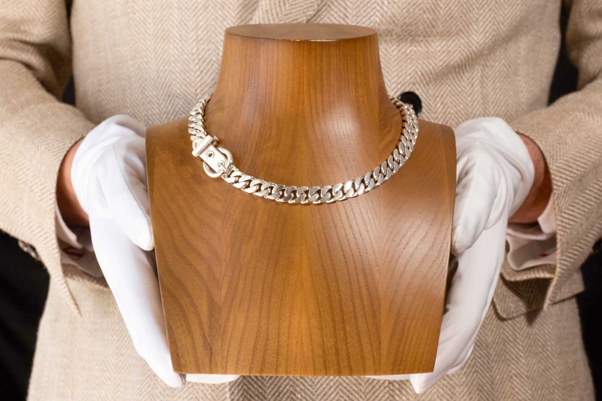 Hermès Ceinture sterling silver necklace on wooden jewelry bust showing belt buckle clasp and anchor chain links against neutral beige and white clothing background