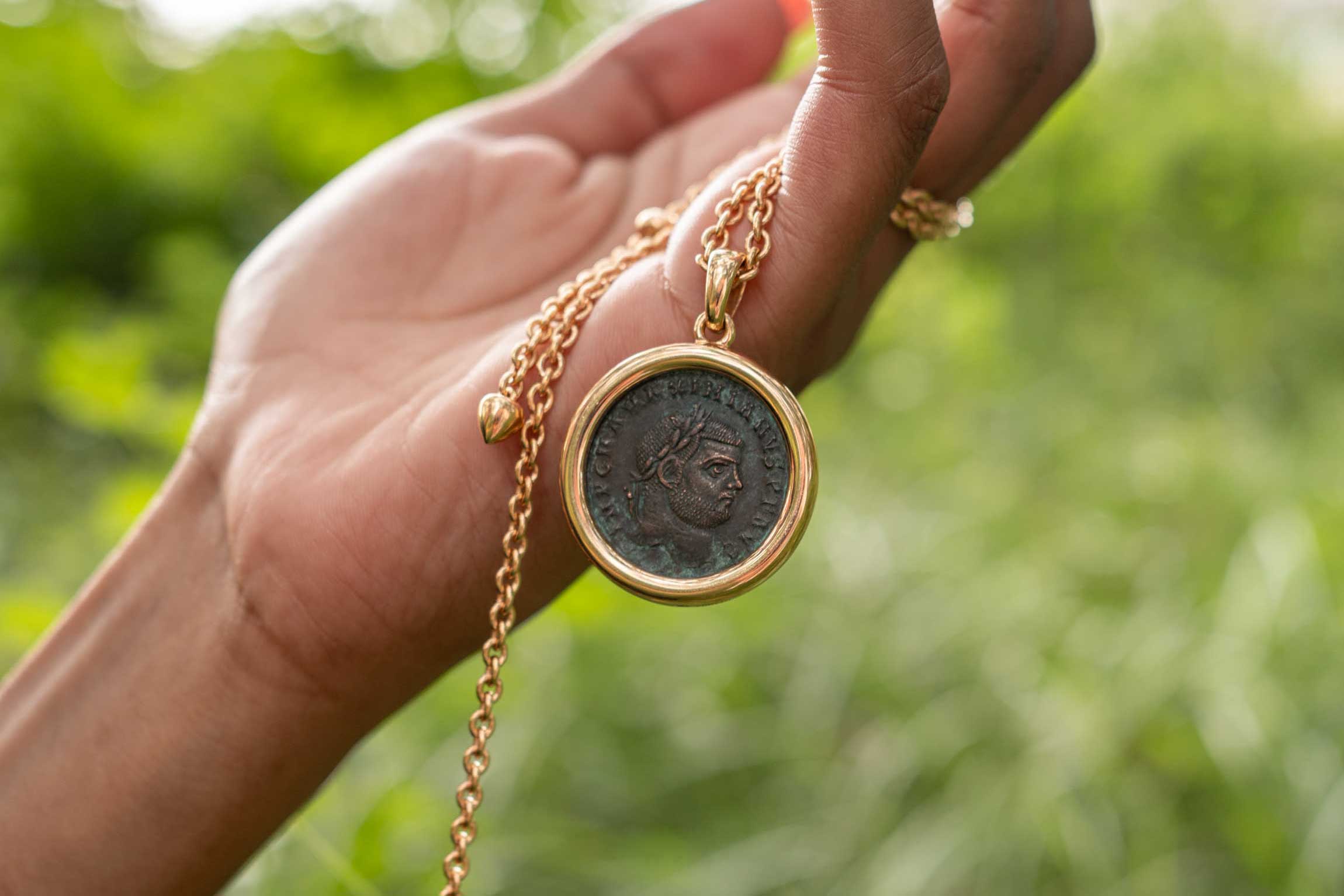A hand holds the Bulgari Monete pendant against a green natural background, showing the ancient bronze Roman coin with its patina and imperial profile set in a polished 18K yellow gold bezel